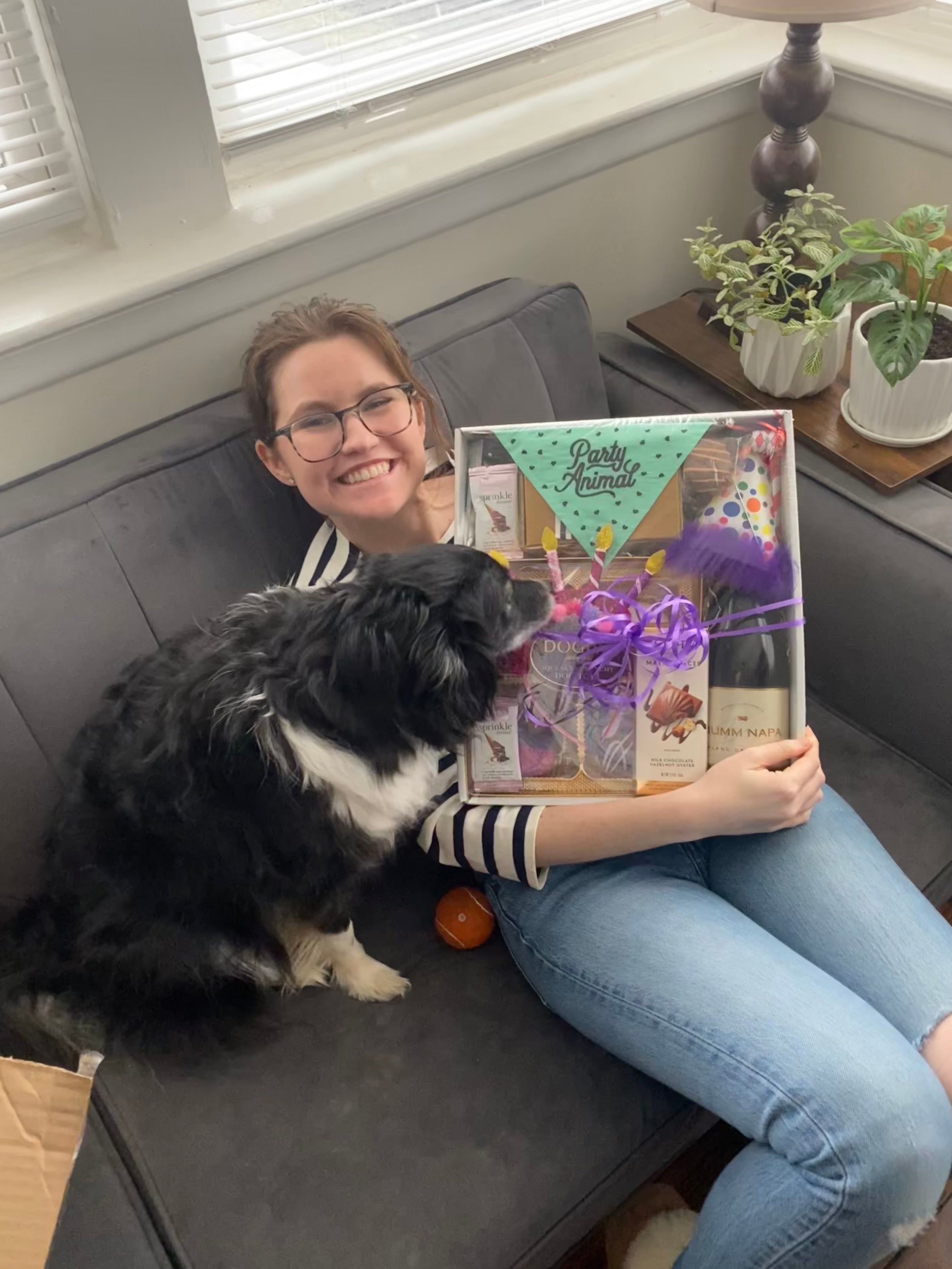 Person sitting on a couch with a dog, holding a gift box, in a home setting.