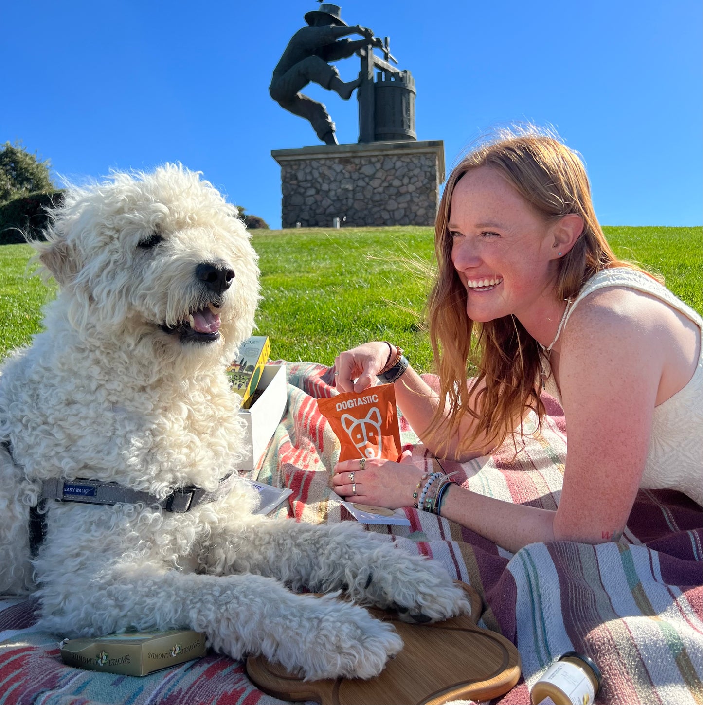 Woman with a white dog sitting on a blanket in a park with a statue in the background enjoying a gift basket by lucca the napa dog
